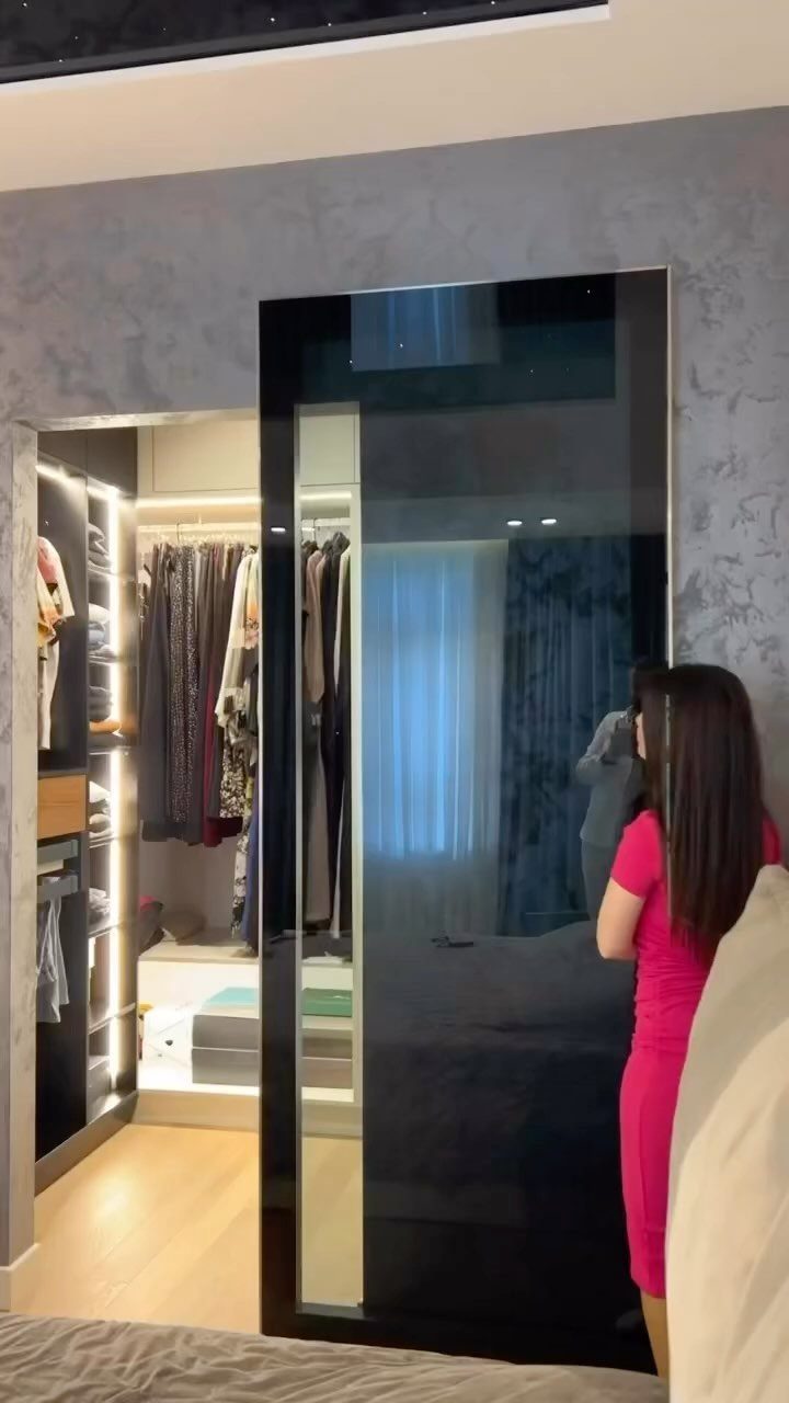 9 Hidden Storage Wall Ideas for a Cleaner Home 3 A large mirror slides open to reveal a well-lit walk-in closet filled with clothing.