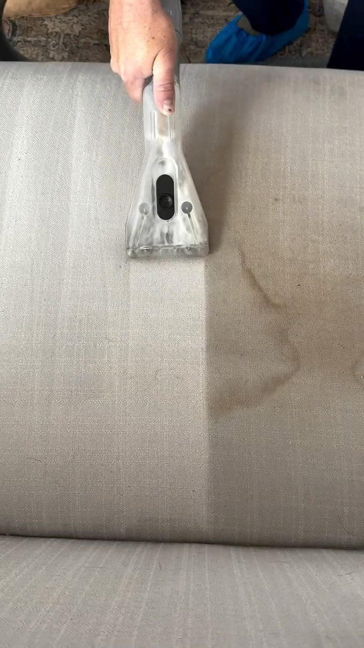A handheld upholstery cleaner removes a large stain from a beige fabric sofa cushion.