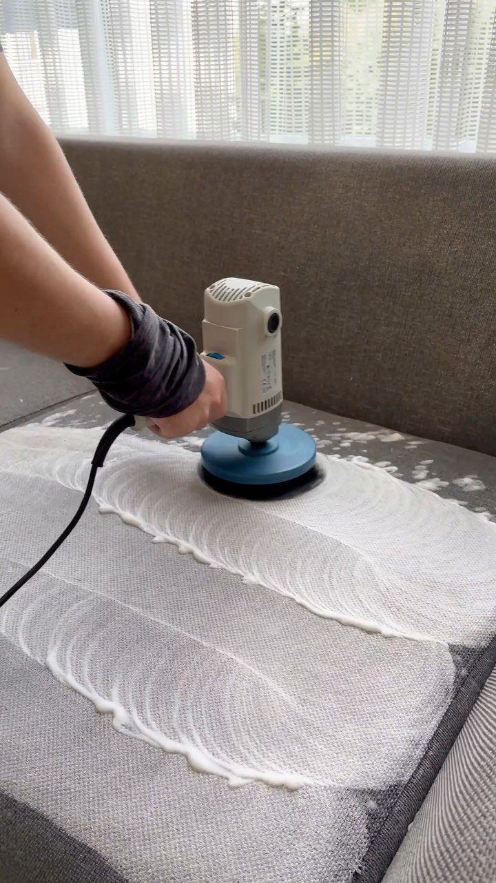 Hands using an electric rotary tool to scrub white cleaning foam onto a grey fabric sofa.