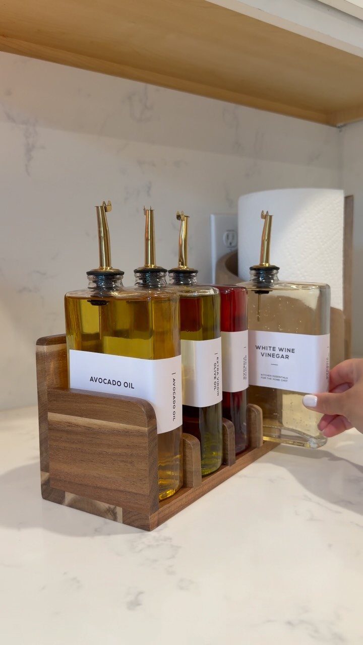 Glass cruets with white labels sit in a wooden holder on a marble countertop.