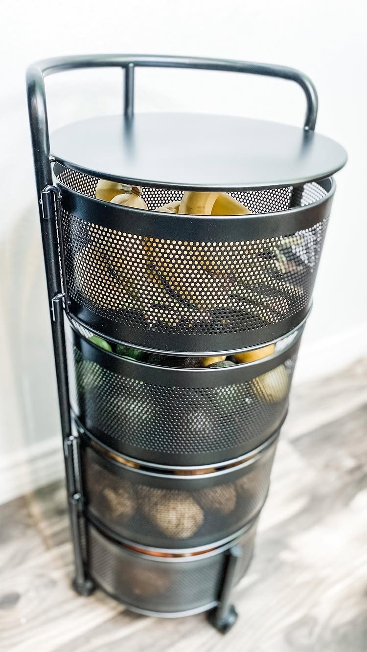 A black metal tiered basket stand filled with various fruits and vegetables on a wooden floor.