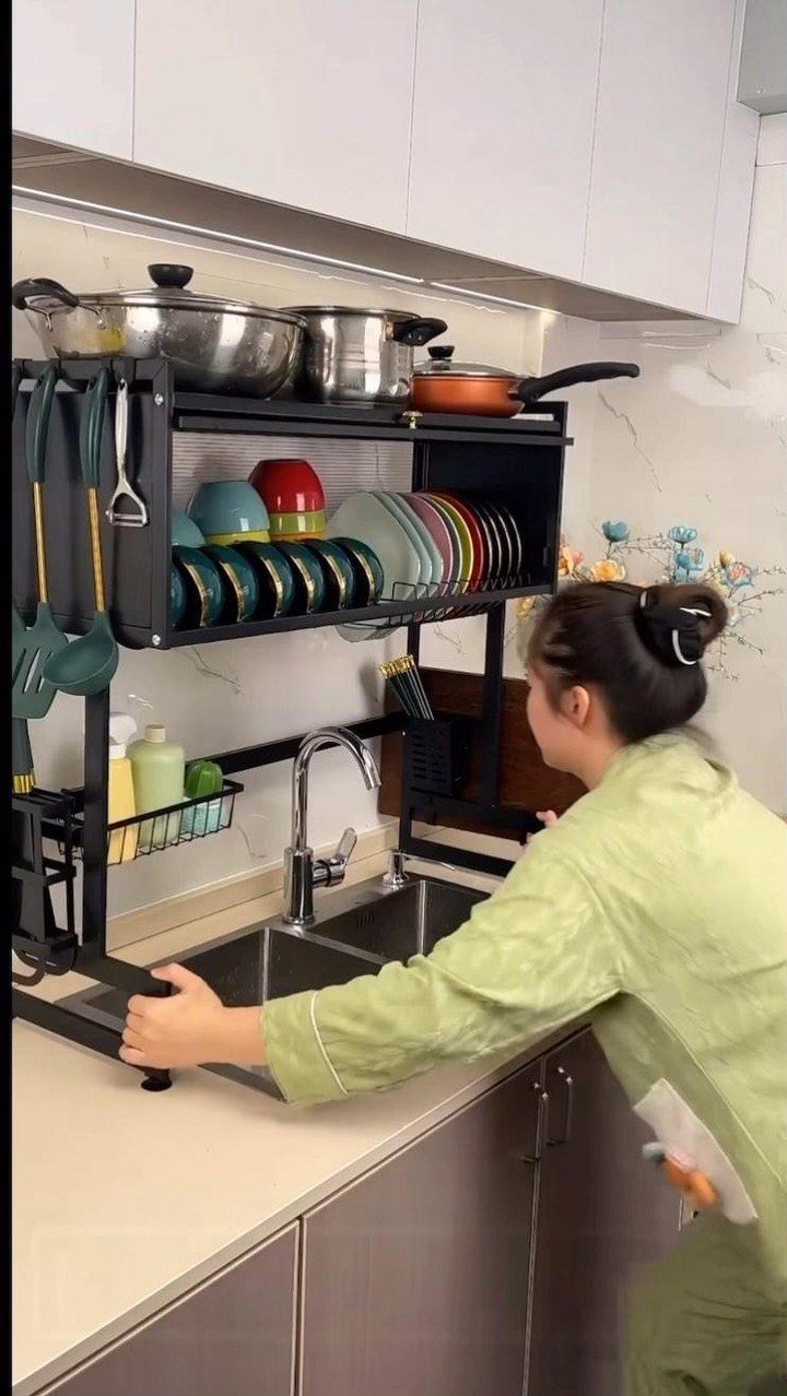 9 Kitchen Tools Organization Ideas for Cabinets, Drawers & Walls 5 A person adjusting a black multi-tier metal rack holding kitchenware over a sink.