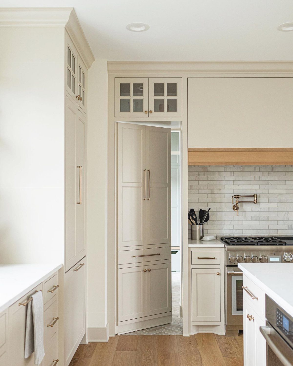 Kitchen with Hidden Pantry: 8 Ideas That Blend Right In 7 Cream kitchen cabinets and a tall paneled pantry door standing slightly open in a bright room.