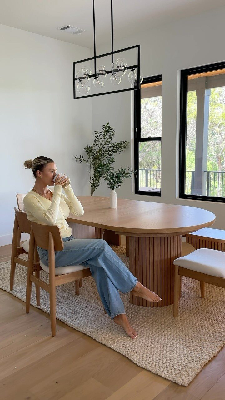 A woman sitting at a large oval wooden dining table in a bright modern dining room.