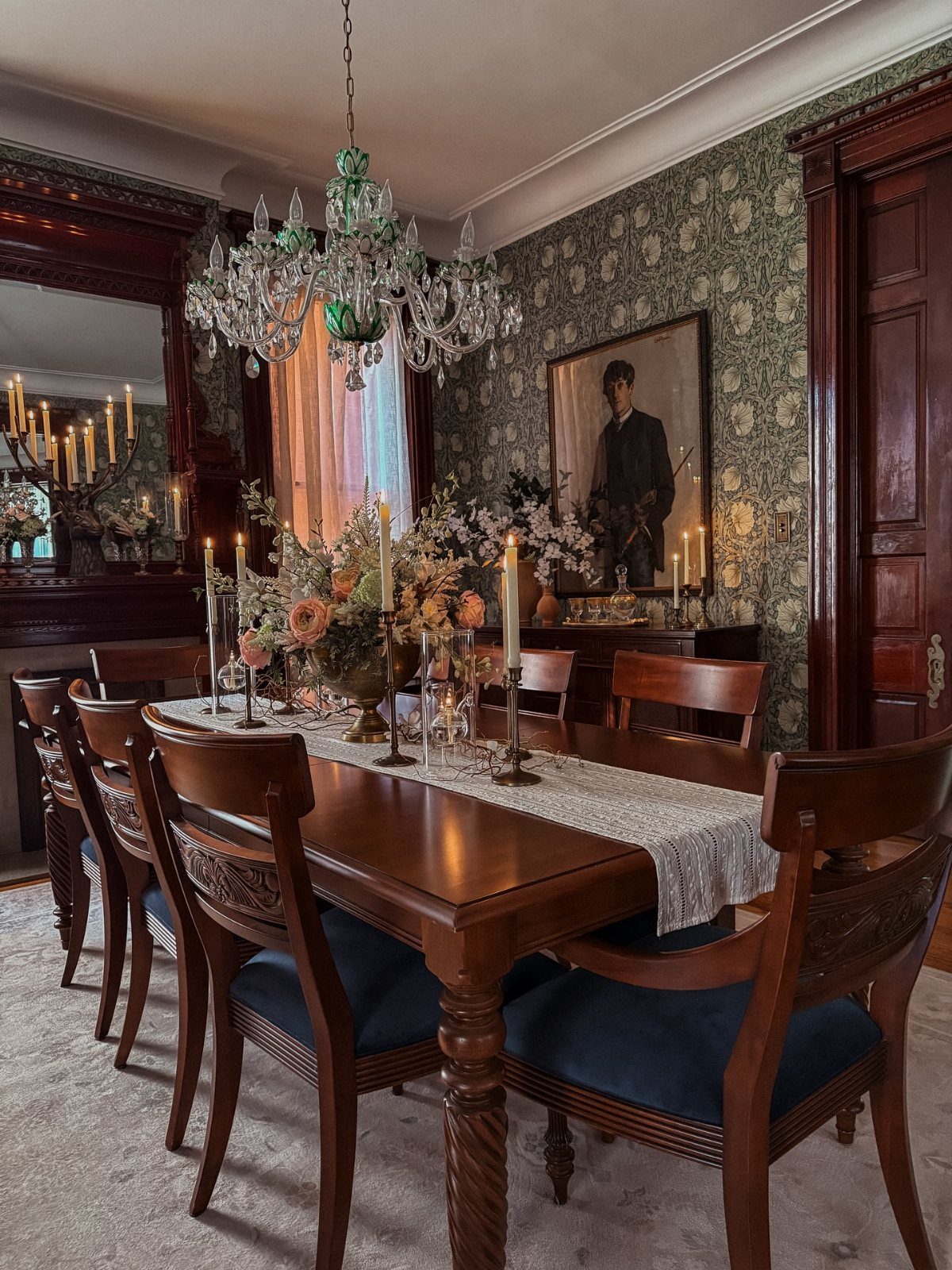 A formal dining room with a wooden table, blue chairs, crystal chandelier, and patterned wallpaper.