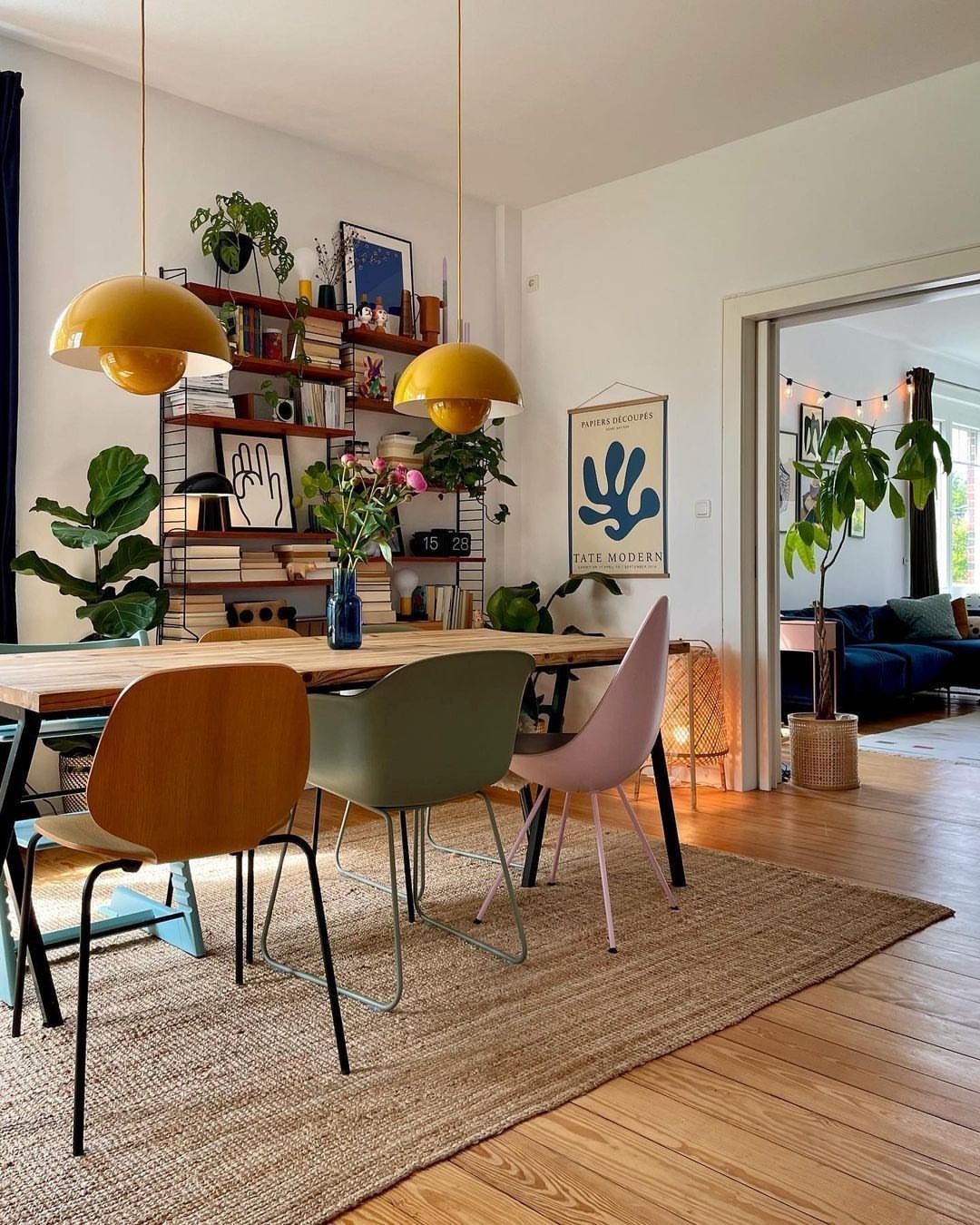 Mismatched dining chairs in wood, green, and pink surrounding a wooden table under yellow pendant lights.