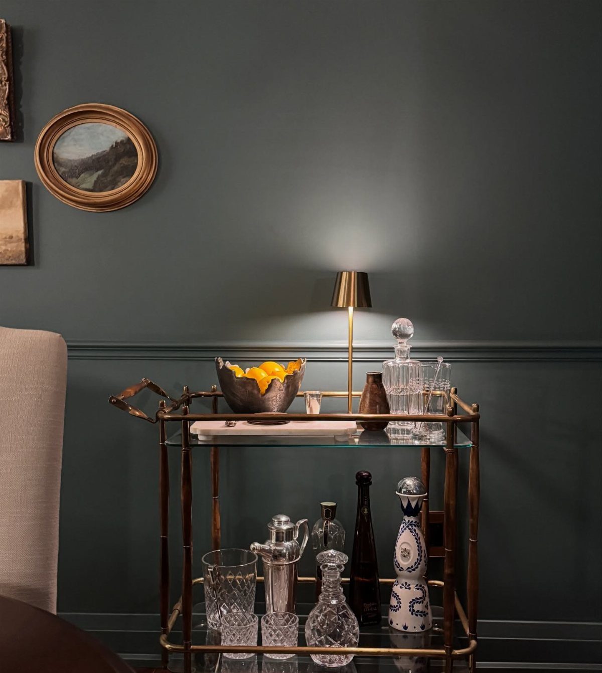 A gold metal bar cart holding various glassware and a small lamp against a dark wall.