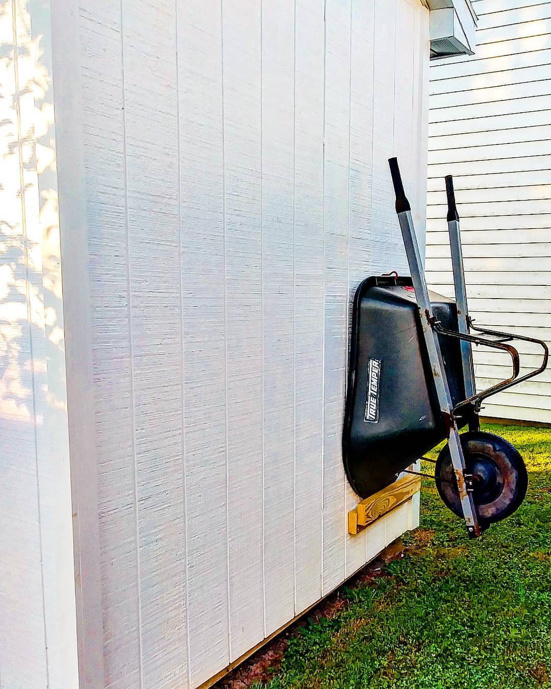 14 Shed Organization Ideas for a Tidy, Functional Space 8 A black wheelbarrow mounted vertically against a white shed wall using a wooden bracket.