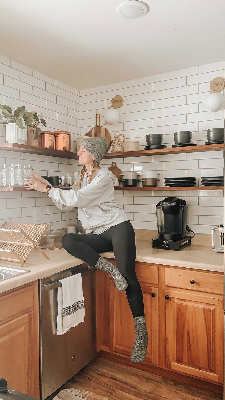 18 Small Kitchen Remodel Ideas to Open Up Your Space 2 A woman sitting on a kitchen counter in front of wooden open shelving and subway tiles.